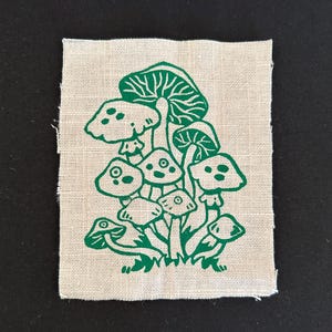 Puede incluir: Un parche cuadrado de tela de color natural con una ilustración verde de varias setas. Las setas tienen sombreros y tallos detallados, algunos con pequeños puntos. El diseño está centrado en la tela.