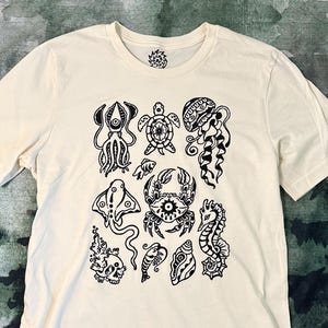 Könnte beinhalten: Cremefarbenes T-Shirt mit schwarzen Illustrationen von Meereskreaturen, darunter ein Oktopus, eine Schildkröte und eine Qualle. Das Shirt hat einen Rundhalsausschnitt und kurze Ärmel. Ein kleines kreisförmiges Design befindet sich auf der oberen linken Brust.