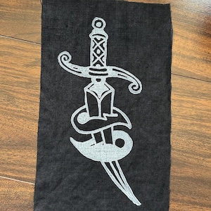 May include: A rectangular black fabric patch featuring a screen-printed design of a silver dagger. The dagger is depicted with a detailed hilt and a blade wrapped in a stylized silver ribbon or banner element, centered on the dark textile surface.