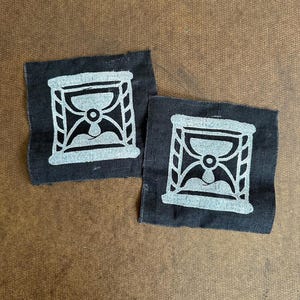 May include: Two square black fabric patches, each featuring a screen-printed white graphic of an hourglass. The design shows the sand flowing from the top bulb to the bottom bulb within a rectangular frame, with textured edges on the fabric.