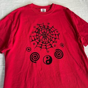 Puede incluir: Una camiseta roja de manga corta con un estampado gráfico negro en el pecho. El diseño incluye una araña en una telaraña, dos formas de espiral, varios círculos pequeños y un símbolo de yin-yang. La camiseta tiene cuello redondo y una etiqueta.