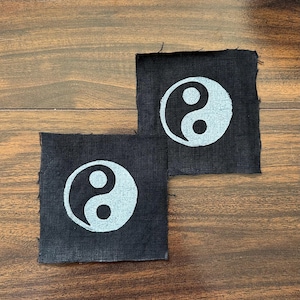 Peut inclure: Deux écussons carrés en tissu noir, chacun arborant un symbole yin et yang sérigraphié en blanc au centre. Les écussons présentent des bords bruts légèrement effilochés et une texture tissée visible, offrant un contraste marqué.