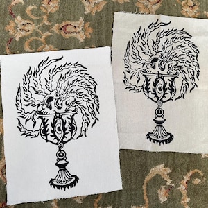 May include: Two rectangular off-white fabric patches featuring a black screen-printed graphic. The design depicts a detailed chalice with a skull resting inside, surrounded by swirling flames that extend outward in a circular pattern.