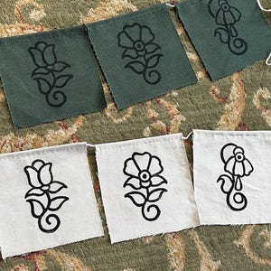 May include: A bunting banner featuring six square fabric flags arranged in two rows. The top row consists of three olive green flags, and the bottom row has three off-white flags. Each flag displays a black line-art illustration of a flower.