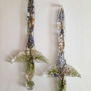 Op de afbeelding: Twee dolken van heldere hars met ingebedde gedroogde bloemen en groen. Elke dolk heeft een puntig blad, een pareerstang en een ronde pommel, met delicate paarse, witte en groene botanische elementen die door het transparante materiaal zichtbaar zijn.