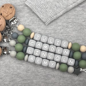 May include: Four wooden teether clips with the text "Little Clips" engraved on each. Each clip is attached to a silicone teething necklace with gray, green, and beige beads. The necklace has the names "JULIE", "CAMDEN", "LINCOLN", and "REMY" spelled out in gray beads.