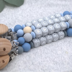 May include: Baby pacifier clips with wooden and silicone beads in shades of blue, gray, and cream. The clips feature names spelled out in lettered blocks. The wooden clips are round with three holes.