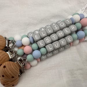 May include: A gray, white, and pastel-colored silicone baby teether with wooden clips. The teether has letter beads that spell out the names "Grayson", "Elliot", "Esther", and "Luke".