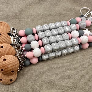 May include: Five wooden teethers with clips attached to a string of gray and pink silicone beads with white letters spelling out the names "Renea", "Jaiden", "Kate", "Ellie", and "Sandra".