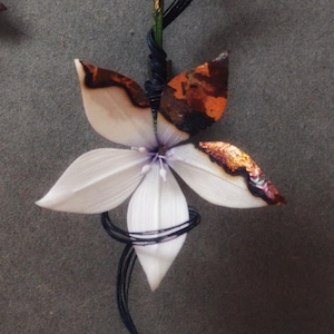 Op de afbeelding: Sieradenset met bloemthema, bestaande uit een paar oorbellen en een hanger. De oorbellen en hanger tonen witte, bruine en zwarte bloemmotieven met druppelvormige kralen. De sieraden zijn geplaatst tegen een grijze achtergrond.