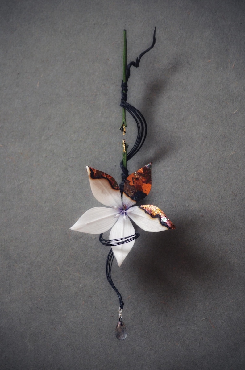Op de afbeelding: Een handgemaakt bloemenornament met witte bloemblaadjes en accenten van bruin en goud. De bloem is bevestigd aan een groene stengel gewikkeld met zwart draad, met een kleine kristallen hanger eronder.