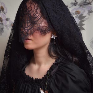 Op de afbeelding: Een persoon draagt een zwarte kanten sluier en een zwarte jurk. Het gedetailleerde patroon van de sluier contrasteert met de jurk. Een bloemenoorbel en een bloemenhanger voegen delicate details toe aan het donkere ensemble. De algehele esthetiek is gotisch en elegant.
