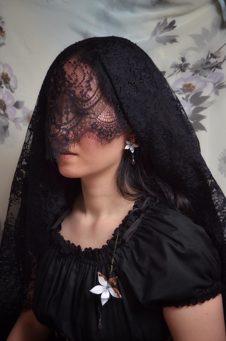 Op de afbeelding: Een persoon draagt een zwarte kanten sluier en een zwarte jurk. Het gedetailleerde patroon van de sluier contrasteert met de jurk. Een bloemenoorbel en een bloemenhanger voegen delicate details toe aan het donkere ensemble. De algehele esthetiek is gotisch en elegant.