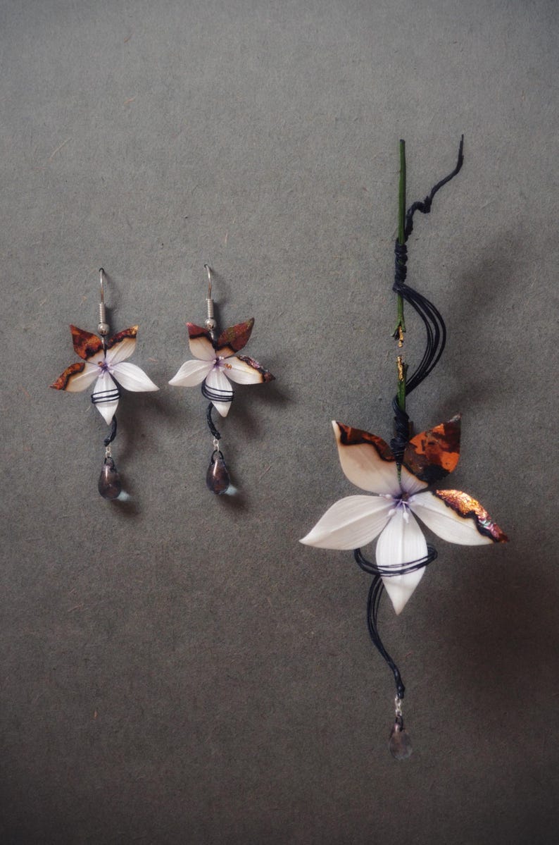 Op de afbeelding: Sieradenset met bloemthema, bestaande uit een paar oorbellen en een hanger. De oorbellen en hanger tonen witte, bruine en zwarte bloemmotieven met druppelvormige kralen. De sieraden zijn geplaatst tegen een grijze achtergrond.