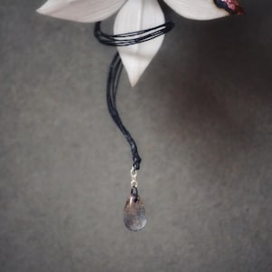 Op de afbeelding: Een druppelvormige hanger hangt aan een zwart koord, onder een witte bloem met een donker centrum en een glinsterende, veelkleurige rand. De hanger is een mix van grijze en bruine tinten, met een zilveren sluiting.