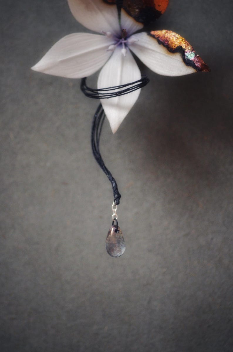 Op de afbeelding: Een druppelvormige hanger hangt aan een zwart koord, onder een witte bloem met een donker centrum en een glinsterende, veelkleurige rand. De hanger is een mix van grijze en bruine tinten, met een zilveren sluiting.