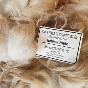 May include: A close-up of a 4 ounce bag of 100% Navajo Churro wool, natural white, from Cedar Mesa Ranch, LLC. in Dolores, Colorado. The wool is a light brown color and is very fluffy.