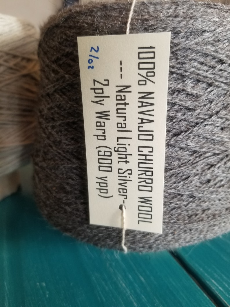May include: A skein of 100% Navajo Churro wool yarn in a light silver color. The label reads "100% Navajo Churro Wool -- Natural Light Silver - 2/02 2ply Warp (900 ypp)".