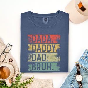 May include: A navy blue t-shirt with the words "DADA. DADDY. DAD. BRUH." in a distressed, retro style. The text is layered in red, yellow, green, and blue. The shirt is folded and displayed with a hat, watch, and glasses.