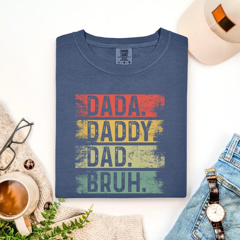 May include: A navy blue t-shirt with the words "DADA. DADDY. DAD. BRUH." in a distressed, retro style. The text is layered in red, yellow, green, and blue. The shirt is folded and displayed with a hat, watch, and glasses.