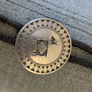 May include: A silver-colored bolo tie clasp with an engraved geometric design and a central image of an animal. The clasp is round and has a black cord. The background is a textured, light green fabric.