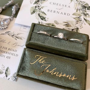 May include: A rectangular olive green velvet ring box with the gold text The Johnsons, containing three silver rings. Nearby are silver teardrop earrings, a white wedding invitation with a green botanical wreath, and a beige box with Mr & Mrs Garcia.