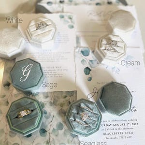 May include: A selection of velvet ring boxes in white, cream, sage, and seaglass hues, each containing a ring. Wedding invitations with eucalyptus leaf patterns are visible. The boxes are octagonal.