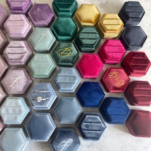 May include: Assortment of hexagonal velvet ring boxes in various colors, including blue, green, purple, red, and gold. Some boxes have initials or the word "Bride" embroidered on the lid.