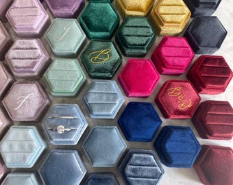 May include: Assortment of hexagonal velvet ring boxes in various colors, including blue, green, purple, red, and gold. Some boxes have initials or the word "Bride" embroidered on the lid.