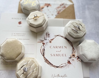 May include: A collection of cream-colored velvet hexagonal ring boxes arranged around wedding stationery. The stationery features a floral wreath illustration, the names Carmen and Samuel, and the date 07.08.24. Several rings are displayed in the boxes.
