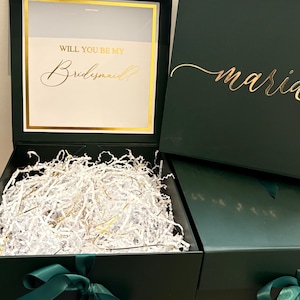 May include: Open green gift box with a gold-framed window displaying the text "WILL YOU BE MY Bridesmaid?". The box is filled with white shredded paper and has a green ribbon. The lid of a second box is visible, with the name "maria" in gold script.