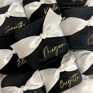 May include: A collection of black velvet jewelry boxes with white satin ribbon bows and gold lettering. The boxes are personalized with names such as "Elise", "Kayla", "Samantha", "Megan", "Brigitte", and "Audrey".