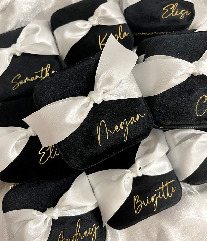 May include: A collection of black velvet jewelry boxes with white satin ribbon bows and gold lettering. The boxes are personalized with names such as "Elise", "Kayla", "Samantha", "Megan", "Brigitte", and "Audrey".
