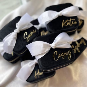 May include: A group of black velvet jewelry boxes with white satin bows and gold lettering. Each box has a different name on it, including Katie, Sami, Casey, and Stephanie.