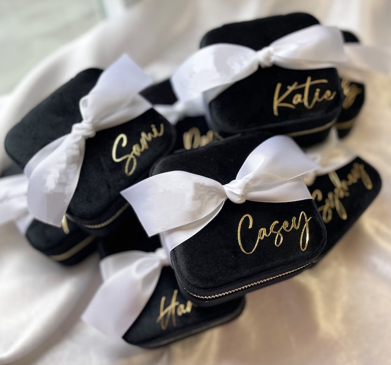 May include: A group of black velvet jewelry boxes with white satin bows and gold lettering. Each box has a different name on it, including Katie, Sami, Casey, and Stephanie.