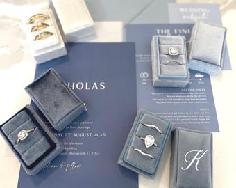 May include: An assortment of engagement rings and wedding bands displayed in gray velvet ring boxes. Wedding invitations and stationery in shades of blue are arranged around the rings. The invitation reads "HOLAS" and "DAY 7TH AUGUST 2026".
