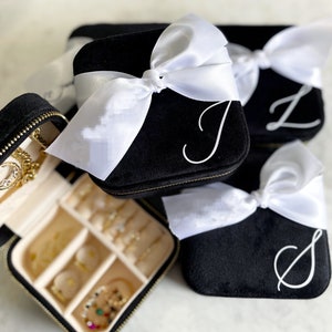 May include: Three black velvet jewelry boxes with white satin bows and personalized initials. The boxes are stacked on top of each other. The top box is open and shows a beige interior with compartments for storing jewelry.