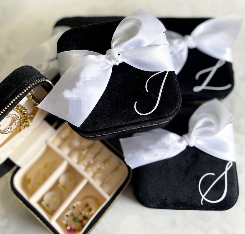 May include: Three black velvet jewelry boxes with white satin bows and personalized initials. The boxes are stacked on top of each other. The top box is open and shows a beige interior with compartments for storing jewelry.