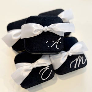 May include: A stack of five black velvet ring boxes with white satin ribbon bows. Each box is personalized with a white letter: A, E, M.