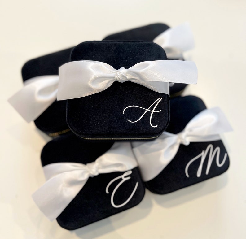 May include: A stack of five black velvet ring boxes with white satin ribbon bows. Each box is personalized with a white letter: A, E, M.
