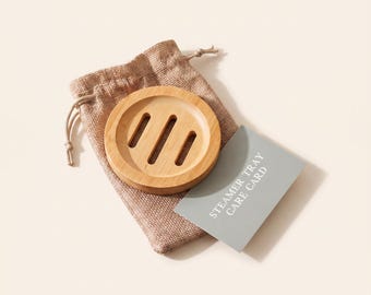 May include: A round light brown bamboo steamer tray with three vertical drainage slots, placed on a tan drawstring burlap pouch next to a grey rectangular card with the white printed text STEAMER TRAY CARE CARD.