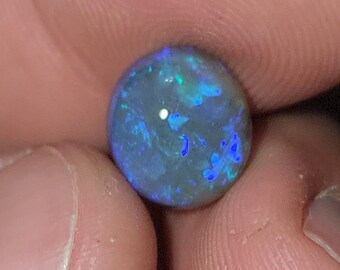 May include: A round, polished opal gemstone with a dark base color and vibrant flashes of blue and green. The gemstone is held between two fingers, showcasing its iridescent play-of-color. The opal appears to be a cabochon cut.