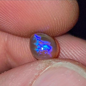 Könnte beinhalten: Ein ovales Opal-Edelstein mit einem durchscheinenden Körper und lebendigen blauen und violetten Farbspielen im Inneren. Der Edelstein wird zwischen zwei Fingern gehalten, was seine irisierenden Eigenschaften hervorhebt. Die Oberfläche des Opals reflektiert das Licht.