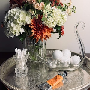 May include: A silver-toned metal tray holding a glass vase with white and orange flowers, a glass swan-shaped dish containing white spherical objects, a small glass holder with cotton swabs, and an orange tube of hand cream.