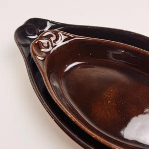 May include: Two nested oval-shaped baking dishes with a glossy dark brown glaze. The smaller dish features a decorative embossed scroll pattern on the handle area. Both pieces have a smooth, reflective finish and a deep, elongated basin design.