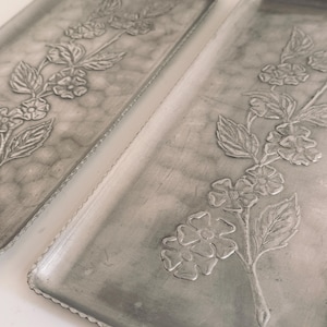May include: Two rectangular silver-toned metal trays featuring a raised floral motif of a flowering branch with leaves and blossoms. The trays have a textured surface and a scalloped edge detail along the perimeter of the metal frame.