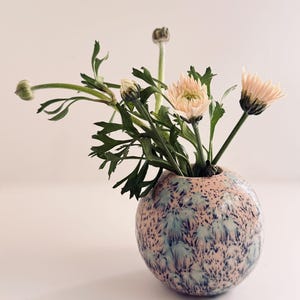 May include: A round ceramic vase with a speckled pink and blue glaze pattern. The vase contains a small bouquet of green stems and light pink flowers, including two fully bloomed flowers and two closed buds, extending from the narrow opening.