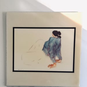 May include: A framed art print featuring a sketch of a figure with dark hair in a bun, wearing a textured blue top. The figure is positioned with one hand on the ground, while the lower body is rendered in simple, light pencil outlines.