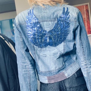 Puede incluir: Una chaqueta vaquera azul con un diseño de alas de ángel de lentejuelas azules en la espalda. La chaqueta tiene una cintura de punto acanalado gris oscuro.