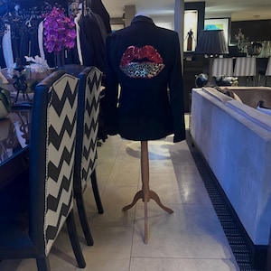 May include: Black blazer displayed on a wooden mannequin. The back of the blazer features a large sequined lips design with red and leopard print accents. The blazer is displayed in a boutique setting with other clothing racks and furniture.